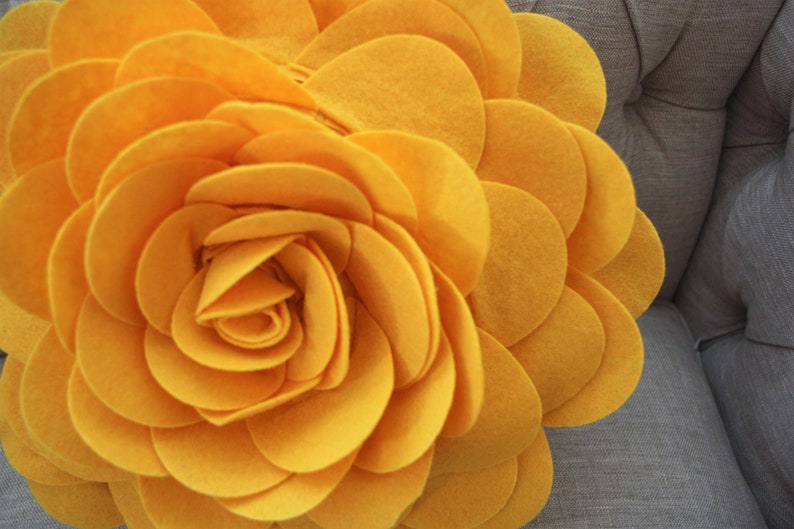 K&ouml;nnte beinhalten: Ein gro&szlig;es, gelbes Kissen in Form einer Filzblume. Die Blume besteht aus vielen Schichten von Filzkreisen, die einen 3D-Effekt erzeugen.