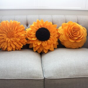 K&ouml;nnte beinhalten: Drei gelbe Filzblumen-Kissen auf einem grauen Sofa. Die Kissen sind in Form von Sonnenblumen und Rosen.