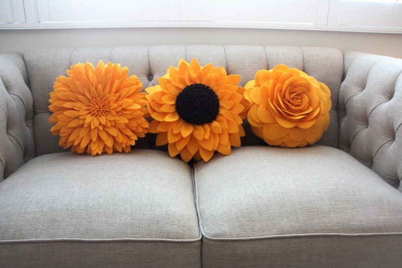 K&ouml;nnte beinhalten: Drei gelbe Filzblumen-Kissen auf einem grauen Sofa. Die Kissen sind in Form von Sonnenblumen und Rosen.