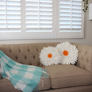 May include: A beige tufted sofa with two white daisy-shaped pillows with orange centers. A light blue and white checkered throw blanket with fringe is draped over the sofa. White shutters are in the background.