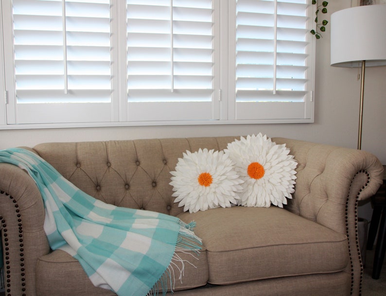 May include: A beige tufted sofa with two white daisy-shaped pillows with orange centers. A light blue and white checkered throw blanket with fringe is draped over the sofa. White shutters are in the background.
