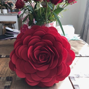 K&ouml;nnte beinhalten: Ein gro&szlig;es rotes Filzrosen-Kissen mit einem geschichteten Design. Das Kissen besteht aus mehreren Schichten Filz, wodurch ein 3D-Effekt entsteht.