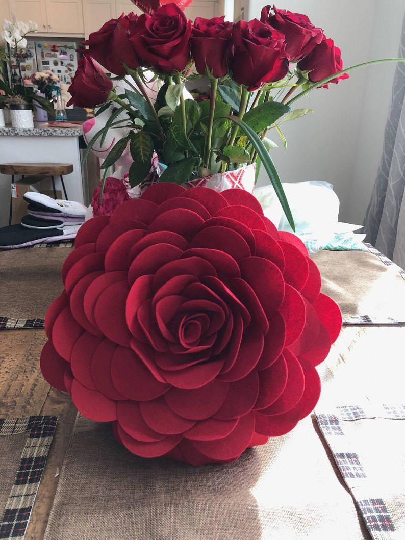 K&ouml;nnte beinhalten: Ein gro&szlig;es rotes Filzrosen-Kissen mit einem geschichteten Design. Das Kissen besteht aus mehreren Schichten Filz, wodurch ein 3D-Effekt entsteht.