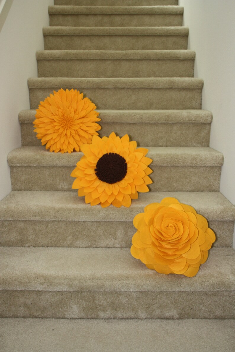 K&ouml;nnte beinhalten: Drei gro&szlig;e gelbe Filzblumen-Dekorationen auf einer mit Teppich ausgelegten Treppe. Die Blumen haben die Form von Sonnenblumen und Rosen.