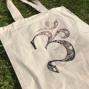 May include: A white canvas tote bag with a black and white graphic of the Om symbol. The symbol is surrounded by a swirling design of stars and moons.