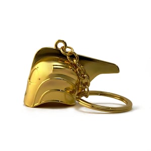 May include: Gold-colored metal keychain featuring a helmet design with a chain and ring.