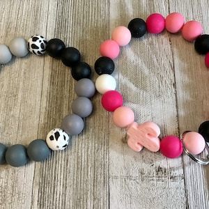 Peut inclure: Deux porte-clés en perles de silicone avec un charme cactus. Un porte-clés est gris, noir et blanc avec une perle à imprimé vache. L'autre porte-clés est rose, noir et blanc avec un charme cactus rose. Les deux porte-clés ont un fermoir en métal argenté.