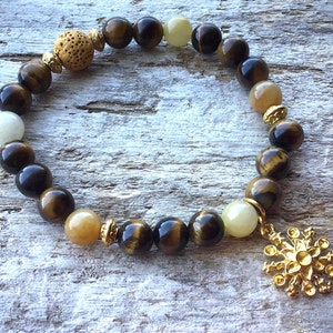 May include: A brown and gold beaded bracelet with a gold charm. The bracelet is made with tiger's eye, moonstone, and lava rock beads.