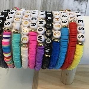 May include: A collection of colorful beaded bracelets. Each bracelet features a different color of flat disc beads and white letter beads spelling out words like "LOVE", "NOW", and "BLESS". The bracelets are stacked together, showcasing a variety of colors and messages.