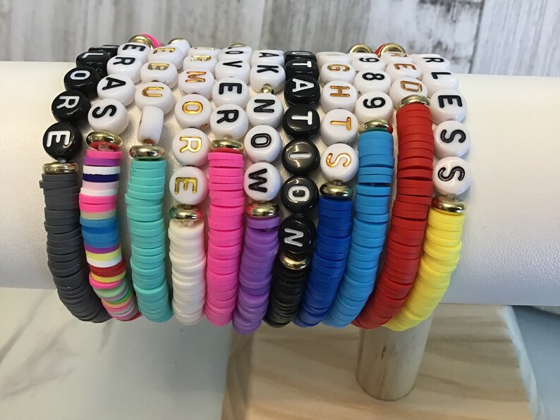 May include: A collection of colorful beaded bracelets. Each bracelet features a different color of flat disc beads and white letter beads spelling out words like "LOVE", "NOW", and "BLESS". The bracelets are stacked together, showcasing a variety of colors and messages.