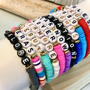 May include: A collection of colorful bracelets with various inspirational words spelled out in gold-lettered beads. The bracelets are made of a mix of black, white, and brightly colored beads. The words include "Explore", "More", and "Fearless".