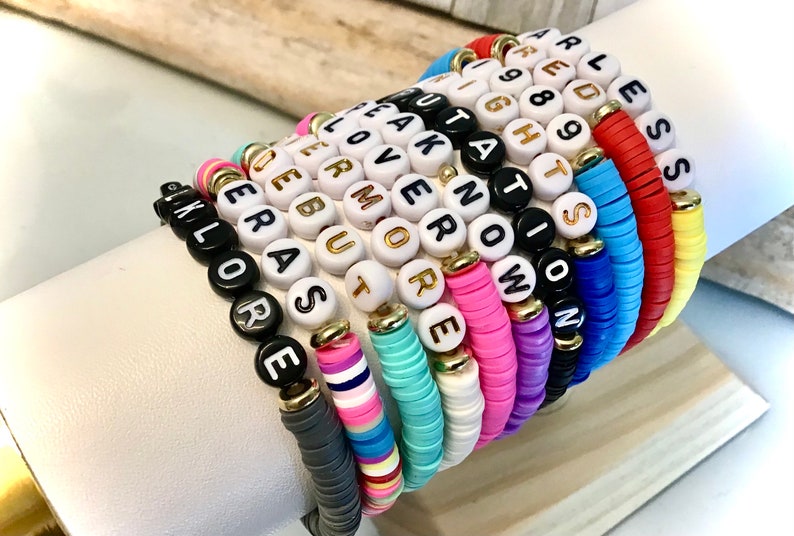 May include: A collection of colorful bracelets with various inspirational words spelled out in gold-lettered beads. The bracelets are made of a mix of black, white, and brightly colored beads. The words include "Explore", "More", and "Fearless".
