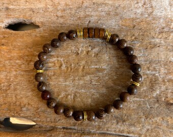 May include: A beaded bracelet featuring dark brown wooden beads with a striped pattern. The bracelet includes gold-colored metal accents and a central section of rectangular wooden beads. The bracelet is displayed on a wooden surface.