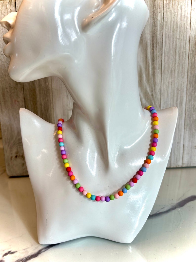 May include: A colorful beaded necklace with a rainbow of colors, including pink, purple, blue, green, yellow, and orange. The necklace is on a white mannequin.