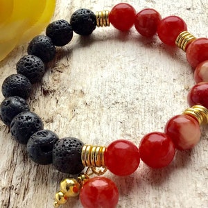 Puede incluir: Un brazalete de cuentas rojas y negras con detalles dorados. El brazalete está hecho con cuentas de roca volcánica y ágata roja.