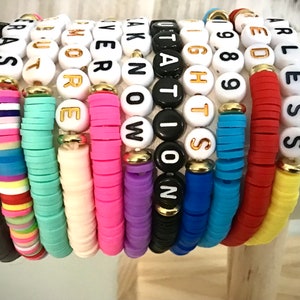 May include: A stack of colorful beaded bracelets. Each bracelet features a different color of flat beads and white letter beads spelling out words. The bracelets are stacked on top of each other, showcasing a variety of colors and phrases.