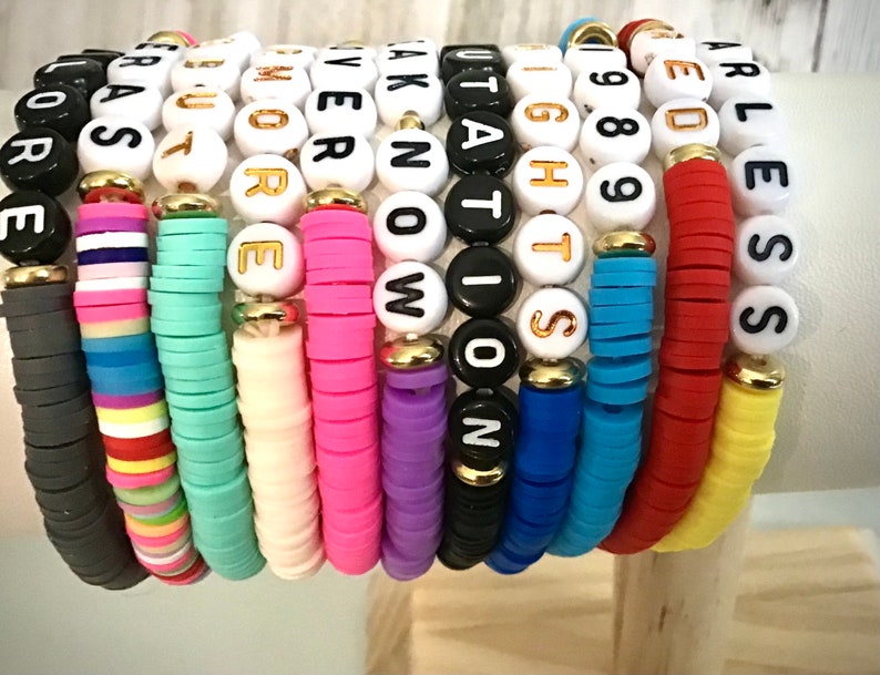 May include: A stack of colorful beaded bracelets. Each bracelet features a different color of flat beads and white letter beads spelling out words. The bracelets are stacked on top of each other, showcasing a variety of colors and phrases.