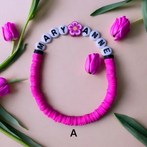 May include: Pink and white beaded bracelet with the name "MARY ANNE" spelled out in black letters. A pink flower charm is in the centre of the bracelet.