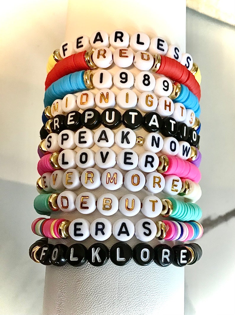 May include: Stack of colorful beaded bracelets with white and black letter beads spelling out words and phrases like "FEARLESS," "RED," "1989," and "FOLKLORE." The bracelets feature gold accents and various colors.