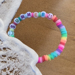 Może przedstawiać: Kolorowa bransoletka z koralików z napisem "SUMMER" napisanym białymi literami. Koraliki są ułożone w tęczę i zawierają zawieszki w kształcie uśmiechniętej buźki.