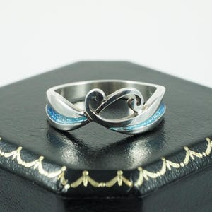 May include: A silver ring with a distinctive design. The ring has a swirling, openwork pattern with blue accents. The blue sections have a glittery appearance, adding a touch of sparkle to the jewellery.