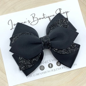 May include: Black glitter hair bow with a layered design. The bow is made of black grosgrain ribbon and black glitter fabric.