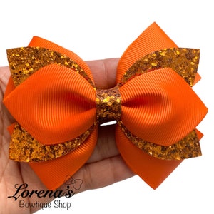 May include: Orange and gold glitter layered hair bow. The bow is made of two layers of orange ribbon with gold glitter trim.