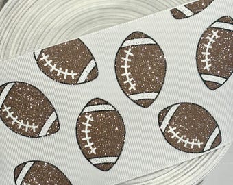 May include: White ribbon with a repeating pattern of brown glitter American footballs, each with white stitching and accents. The ribbon is rolled, displaying its texture and layers. Suitable for crafting and sports-themed projects.