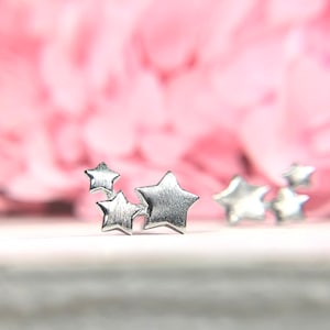 May include: Four silver star-shaped earrings on a white surface. The earrings are small and delicate. The background is blurred and pink.