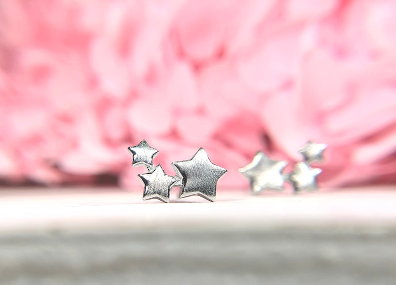 May include: Four silver star-shaped earrings on a white surface. The earrings are small and delicate. The background is blurred and pink.