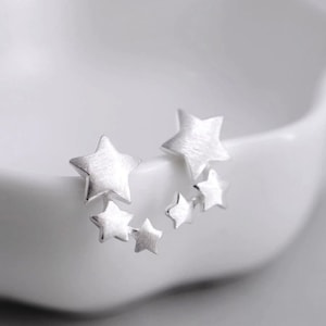 May include: A pair of silver star shaped earrings. Each earring has five stars of varying sizes. The stars are brushed silver and have a matte finish.