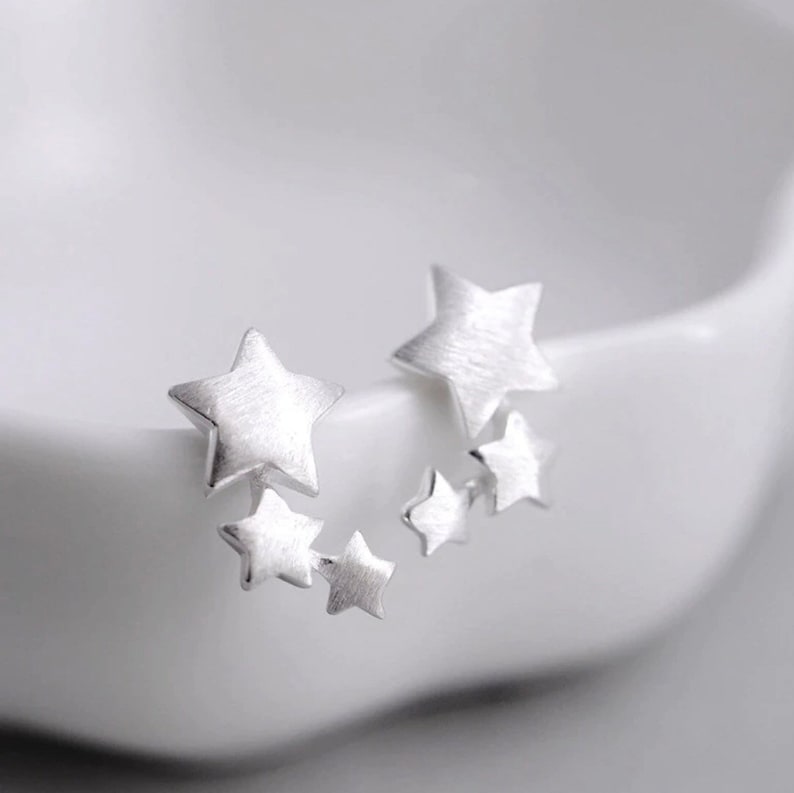 May include: A pair of silver star shaped earrings. Each earring has five stars of varying sizes. The stars are brushed silver and have a matte finish.