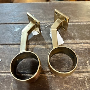 Puede incluir: Dos soportes de pasamanos de metal dorado con base cuadrada y parte superior redonda. Los soportes están diseñados para montarse en una pared o poste y se utilizan para sostener un pasamanos.