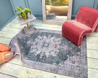 May include: Miniature dollhouse scene featuring a gray and pink patterned rug, a red velvet chair, a decorative mirror, and a small table with a plant. The mirror has a cream-colored frame with floral details. The scene is set against a blue wall.