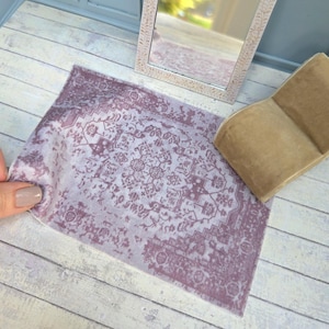 May include: Miniature dollhouse decor featuring a decorative mirror with an ornate, light-colored frame and a rectangular, patterned rug in shades of purple and white. A small, tan chair is also present, suggesting a room setting.