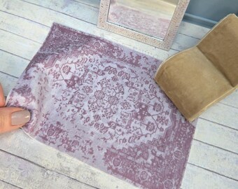 May include: Miniature dollhouse decor featuring a decorative mirror with an ornate, light-colored frame and a rectangular, patterned rug in shades of purple and white. A small, tan chair is also present, suggesting a room setting.