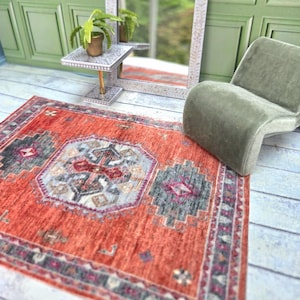 May include: A miniature room setting featuring a decorative mirror with a white floral design, a small side table, a green chair, and a red patterned rug. The room has green paneling on the wall and a light blue floor.