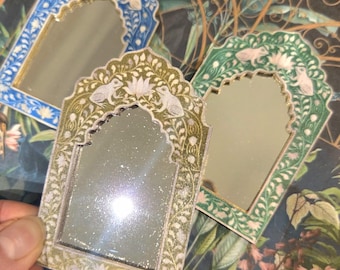 May include: Three decorative mirrors with arched frames. The mirrors have intricate floral and bird designs in blue, green, and gold. The mirrors are held in a hand, and the background has a floral pattern.