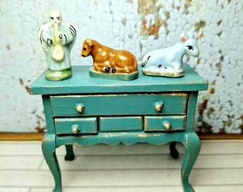 May include: A teal blue dresser with three drawers and gold accents. The dresser has cabriole legs and sits on a white wood floor. Three small figurines are on top of the dresser: a blue and white angel playing a saxophone, a brown calf, and a blue lamb.