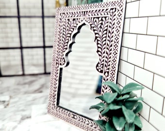 May include: A rectangular mirror featuring a dark brown frame with intricate white inlaid leaf and floral patterns. The mirror has a distinct arched top design. The frame is decorated with a repeating geometric border along the outer edge.