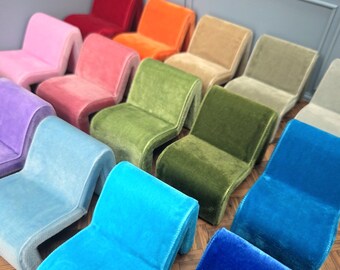May include: A collection of colorful, miniature velvet chairs in various shades, including red, orange, pink, green, blue, and purple. The chairs have a curved, modern design and are arranged in a grid pattern, showcasing their vibrant hues.