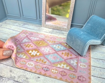 May include: Miniature dollhouse scene featuring a pink and yellow patterned rug, a light blue chair, and a decorative mirror with a floral design. The mirror is framed in a cream color. The rug is approximately 4 inches long.