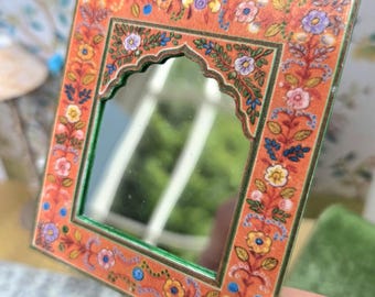 May include: A small, square mirror with an orange floral border. The frame is adorned with colorful flowers and green accents. The mirror's arched top adds a unique touch to this decorative piece.
