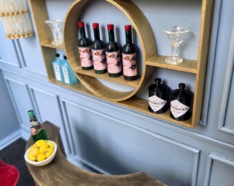 May include: Miniature bar scene with a gold-colored shelf and bar. The shelf holds small bottles of wine, gin, and other beverages, along with cocktail glasses. A small bar with a bowl of lemons and a bottle of Perrier is in the foreground.