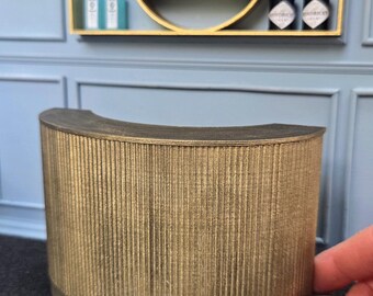 May include: A miniature gold-colored bar with a curved front and vertical ribbed detailing. The bar is set against a blue wall with a gold-framed shelf holding miniature liquor bottles and glasses. The scene evokes a sense of a small-scale, stylish bar.