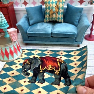 Puede incluir: Alfombra en miniatura con temática de circo con una ilustración de elefante, una manta roja y dorada y un patrón de diamantes azul y crema con estrellas rojas. Un sofá de terciopelo azul y un cojín de jirafa en el fondo.