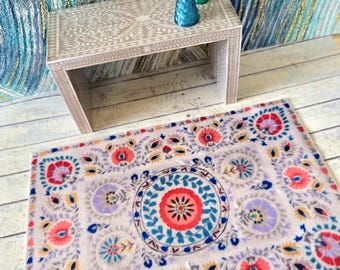 May include: A rectangular area rug featuring a central circular medallion surrounded by floral motifs in shades of coral, blue, and lavender on a light gray background. The rug is positioned on a white wooden floor in front of a matching console table.