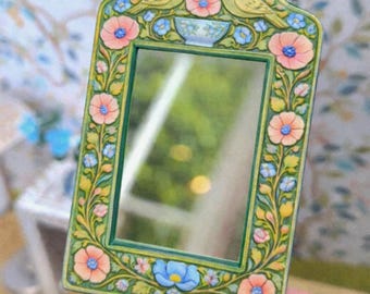 May include: A small, rectangular mirror with a decorative frame. The frame is painted green and adorned with pink and blue flowers, yellow birds, and a blue bowl. The mirror reflects a blurred outdoor scene.