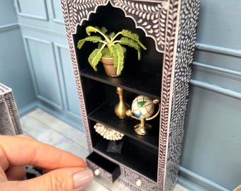 May include: Miniature black and white bookshelf with decorative floral inlay. The shelves hold a potted plant, a gold vase, a globe, and a decorative object. A small drawer is open. The bookshelf is in a dollhouse setting.