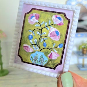 May include: A framed painting of a floral arrangement with pink and blue flowers in a blue vase with two green birds perched on a branch. The painting is set against a green background and has a white frame with a beaded border.